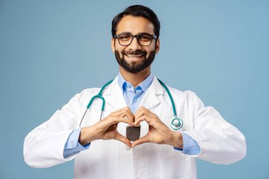 Gülümseyen, mutlu sakallı Hintli bir adamın portresi, şık gözlükler takan bir doktor, beyaz önlük, kalp pili takan, mavi arka planda tek başına duran kameraya bakan bir doktor. Kardiyoloji kavramı