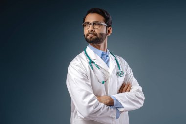 Ciddi, düşünceli Hintli bir adam, beyaz önlüklü bir doktor, steteskop takmış, gözlüklü, kollarını çapraz uzatmış, mavi arka planda tek başına duran. Reklam kavramı