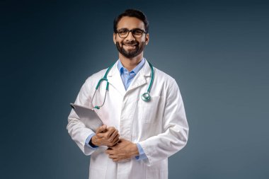 Mutlu, profesyonel Hintli bir adamın portresi, doktor, beyaz ceket, steteskop, dizüstü bilgisayar, kameraya bakıyor, mavi arka planda tek başına duruyor. Tıbbi, reklam konsepti
