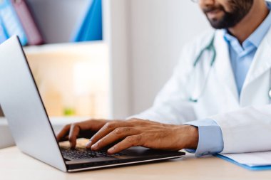 Yakın plan, beyaz önlüklü profesyonel Hintli doktor, klavyeye yazı yazıyor, dizüstü bilgisayar kullanıyor, ellere odaklanıyor, modern klinikte iş yerinde oturuyor. Çevrimiçi teknoloji kavramı