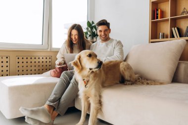Genç çift, modern dairede Golden Retriever köpekleri ile koltukta dinlenirken gülüyor, dizüstü bilgisayar ve telefon kullanıyor.
