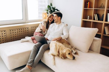 Genç çift evlerinde Golden Retriever köpekleri ile kanepede otururken dizüstü bilgisayar kullanıyor ve gülüyorlar.