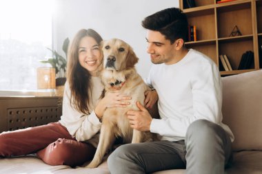 Gülümseyen çift, modern oturma odalarında rahat bir kanepede dinlenirken Golden Retriever köpeğini kucaklıyor.