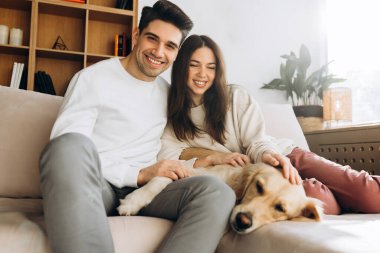 Gülümseyen adam ve kadın evcil köpeğe sarılırken rahat bir oturma odasında rahat bir kanepede oturup hafta sonları evde boş vakitlerin tadını çıkarıyorlar.