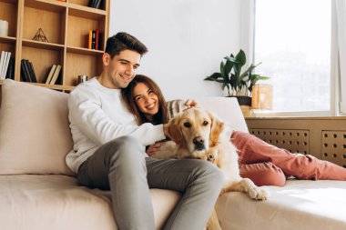 Genç çift rahat bir kanepede otururken kucaklaşıp gülümsüyor ve sevimli Golden Retriever köpekleri rahat bir oturma odasında.