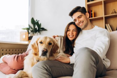 Sevgi dolu genç bir çift rahat bir kanepede tatlı Golden Retriever köpekleri ile gülüyor ve rahatlıyor. Evde huzurlu bir anın tadını çıkarıyorlar.