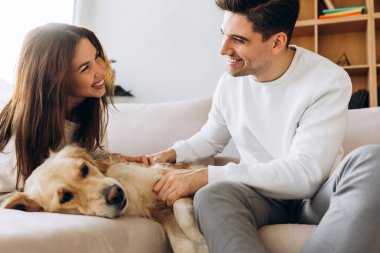 Genç çift, oturma odasındaki rahat bir kanepede dinlenirken Golden Retriever köpeklerini okşayarak birlikte kaliteli zaman geçiriyorlar.