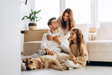 Gülümseyen aile, Golden Retriever köpeğiyle birlikte oturma odasında vakit geçirir. Mutluluk ve aşk anlarının tadını çıkarırlar.