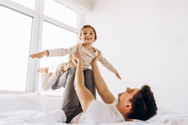 Baba, parlak bir yatak odasında yatakta uçak oyunu oynarken kızını onun üzerine kaldırdı.