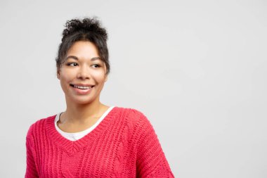 Kıvırcık saçlı, beyaz arka planda pembe bir kazak giyen, mutluluk ve pozitifliği ifade eden neşeli bir genç kadının stüdyo portresi.