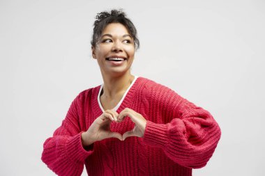 Elleriyle kalp şekli oluşturan ve beyaz bir arka planda izole edilmiş, gülümseyen neşeli genç bir kadının portresi.