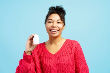 Elinde beyaz plastik bir hap şişesi, vitamin ya da takviye malzemesi tutan mutlu genç bir kadın mavi arka planda gülümsüyor.