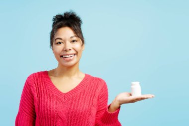 Mavi arka planda beyaz plastik ilaç şişesini sunan neşeli genç bir kadının stüdyo portresi.