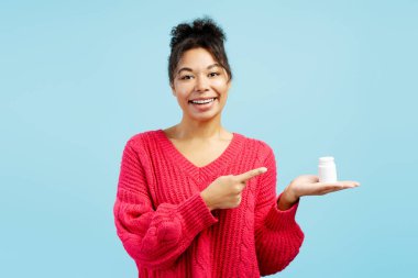 Neşeli genç bir kadın beyaz bir ilaç şişesini işaret ederek tıbbi tedavi veya vitamin öneriyor.