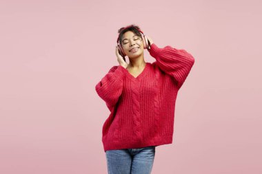 Neşeli genç bir kadın kulaklık takıyor ve rahat bir süveter giyiyor. Yumuşak pembe bir arka plana karşı gevşek bir ifadeyle müzikten hoşlanıyor.