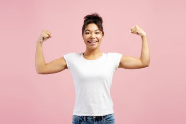 Kaslarını esneten ve gülümseyen pembe bir arka planda gücünü ve güvenini gösteren neşeli genç bir kadının stüdyo çekimi.