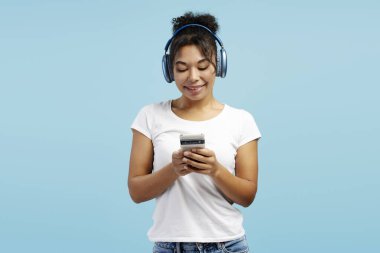 Mavi kulaklık takan gülümseyen kadın, canlı mavi bir arka planda izole haldeyken cep telefonu uygulamasının tadını çıkarıyor.