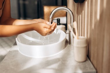 Kadın ellerini, modern ve minimalist bir banyoda temiz suyla yıkıyor. Hijyen ve temizliği vurguluyor.