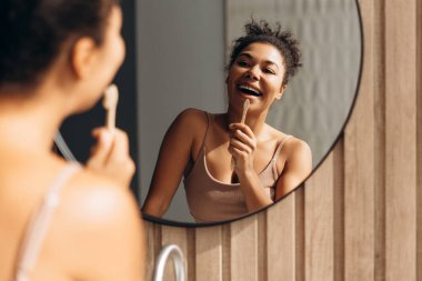 Dişlerini tahta bir diş fırçasıyla fırçalayan mutlu genç bir kadın modern bir banyoda sabah rutininin tadını çıkarıyor.