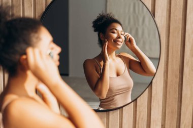 Gülümseyen genç bir kadın yüzüne gua sha taşıyla masaj yapıyor, evdeki sabah güzellik rutininin tadını çıkarıyor.