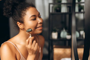 Yüzüne yeşim bir silindirle masaj yapan genç bir kadın, tuvaletinde kendine bakmanın keyfini çıkarıyor.