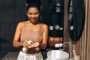 Gülümseyen kadın eline nemlendirici losyon ya da krem sürüyor. Elinde lavabo ve yuvarlak aynayla şık bir banyoda duruyor.