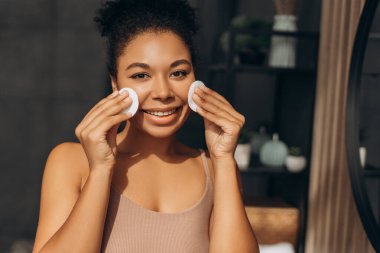 Cilt bakımı için pamuk pedleri kullanan mutlu genç bir kadın. Banyosunda sağlıklı ve temiz bir cildin keyfini çıkarıyor.