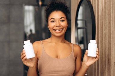 Gülümseyen Afrikalı Amerikalı kadın, parlak bir banyoda iki beyaz plastik takviye şişesi tutuyor. Sağlık ve sağlığın somutlaşmış hali.