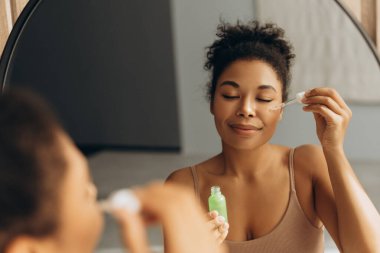 Mutlu melez kadın cilt bakımı için doğal kozmetik serumu kullanıyor. Aynanın önünde sabah rutininin tadını çıkarıyor.