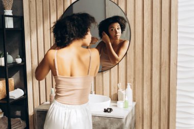 Genç kadın, yuvarlak aynanın önünde, ahşap duvarları olan modern bir banyoda kıvırcık saçlarını şekillendiriyor.