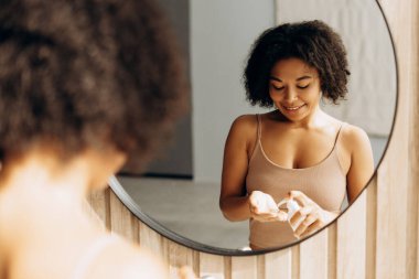 Ellerine nemlendirici sürerken gülümseyen, banyo aynasına yansıyan, kendine bakım ve cilt bakımını kucaklayan genç bir kadın.