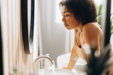 Genç bir kadın banyo lavabosunun üzerine eğiliyor, yüzünü tatlı suyla yıkıyor, ferahlatıcı sabah cilt bakımının keyfini çıkarıyor.