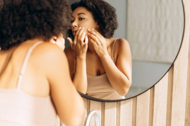 Kıvırcık saçlı kadın banyoda aynaya bakarken yüzünü sıkıyor.