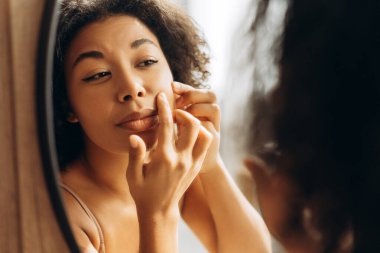 Kıvırcık saçlı genç bir kadın. Evinde aynaya bakarken yüzünde sivilce çıkıyor.