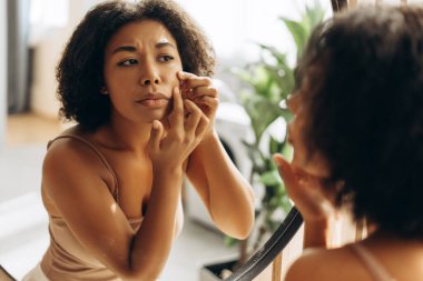 Üzgün bir kadın, banyo aynasında yansımasına bakarken yanağını sıkarak evdeki cilt kusurlarıyla uğraşıyor.