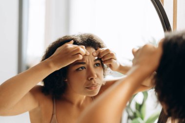 Genç bir kadın, alnına sivilce sıkarken aynadaki yansımasını inceliyor. Banyoda cilt bakımına odaklanıyor.