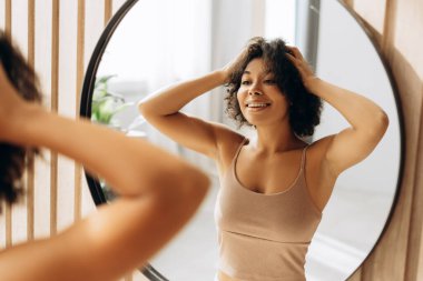 Gülümseyen kadın, yuvarlak aynaya bakarken kıvırcık saçlarını düzeltiyor. Sıcak banyosunda kendine bakmanın keyfini çıkarıyor.