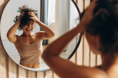 Genç bir kadın aynaya bakarken kıvırcık saçlarını şekillendiriyor, rahat bir banyoda sabah rutininin tadını çıkarıyor.