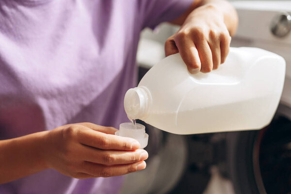 Close up of hands pouring liquid laundry detergent from a white plastic bottle into a measuring cup, preparing for washing clothes