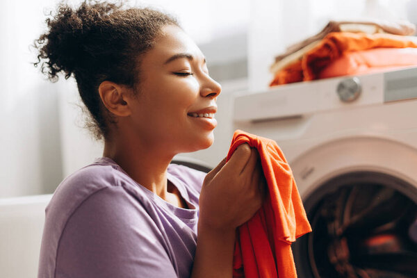 Young woman smelling clean orange clothes after laundry, enjoying fresh scent of detergent and fabric softener