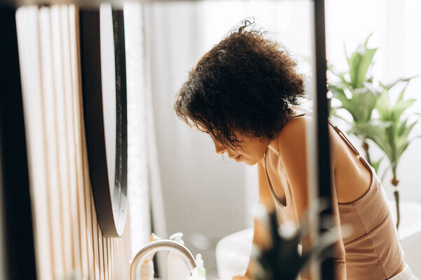 Young woman with curly hair washing her face in a modern and minimalist bathroom, enjoying a moment of self care