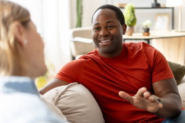 Afro-Amerikan bir adamın, evdeki oturma odasında kanepede oturan bir kadınla konuştuğu portre. Aile eğlencesi, ilişki konsepti