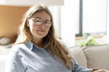 Gözlüklü, gülümseyen ve kameraya poz veren, oturma odasında oturan güzel bir kadın. Yurtiçi yaşam tarzı kavramı