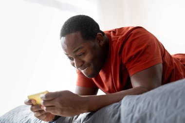 Orta yaşlı mutlu Afro-Amerikan bir adamın portresi. Elinde akıllı telefon, yatak odasında dinleniyor. Teknoloji konsepti