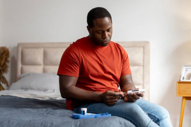 Afro-Amerikalı adam evdeki yatakta oturmuş kan şekerini dijital glikoz sayacıyla ölçüyor. Sağlık hizmeti, tıp konsepti