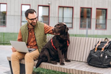 Gülümseyen, mutlu İspanyol bir adamın portresi, dizüstü bilgisayar kullanan serbest çalışan, güzel siyah bir köpeği okşayan, labrador, sokakta, açık havada bankta oturan. Hayvan, evcil hayvan aşkı konsepti
