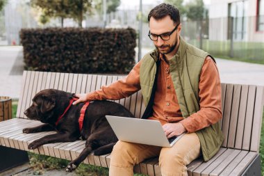 Ciddi orta yaşlı, İspanyol bir adam, günlük kıyafetler giyen serbest çalışan, gözlüklü kısa mesaj atan, dizüstü bilgisayar kullanan, siyah köpeği okşayan, sokakta birlikte oturan. Çevrimiçi iş kavramı