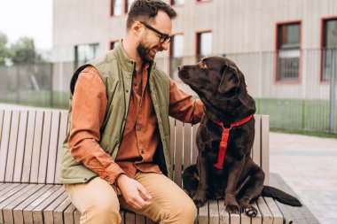 Gülümseyen, orta yaşlı, yakışıklı İspanyol bir adam, siyah köpeği okşuyor, labrador, dışarıda bankta yan yana oturuyor. Hayvan, evcil hayvan aşkı konsepti