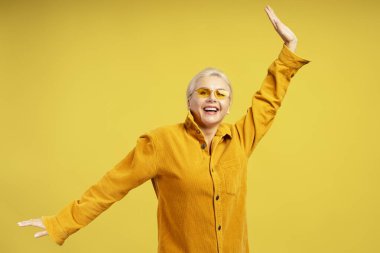 Sarı gömlek ve güneş gözlüğü takan zarif orta yaşlı kadın sarı bir stüdyo arka planında dans ediyor ve eğleniyor.