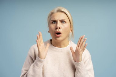 Şaşırmış yetişkin bir kadının stüdyo portresi. Mavi arka planda şaşkınlık ve şaşkınlık içinde ellerini kaldırıyor.